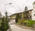 The image shows a residential neighborhood in a mountainous area. There are several multi-story apartment buildings, trees, and a winding road with parked cars. The buildings have balconies visible, and the surrounding landscape includes hills and forests