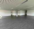 Large open space with gray wood-like flooring, white walls, and a patterned ceiling with recessed lighting. The space appears to be a commercial or office area ready for use or renovation.