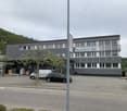 A multi-story commercial building with a large parking lot in front. The building has a grey corrugated metal exterior and several windows. There is a tree in front of the building and a few parked cars in the lot.