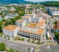 Aerial view of urban buildings, a mix of white buildings with green roofs, a prominent building with red roofs and windows. The surroundings include mountains, greenery, and a busy road network. Numerous vehicles are seen moving along the roads, indicatin