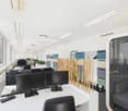 The image shows an open office space with a modern and minimalist design. The room has white walls and a white ceiling with recessed lighting. There are several desks with computers and monitors, as well as storage cabinets and shelves along the walls. Th