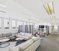 The image shows a modern and open office space with a lounge area. The space features comfortable sofas and armchairs, coffee tables, and a TV mounted on the wall. The ceiling has recessed lighting fixtures and the walls are lined with windows, creating a