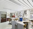 white tiled walls, wooden chairs, desks with tools, sink, cabinets, windows with blinds, bucket, framed picture