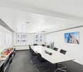 A rectangular office room with white walls, black chairs, a long white table, grey carpet, two large windows, picture and objects on the table