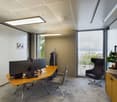 Modern office space with large windows, providing a view. The room features a wooden desk with a computer setup, chairs, and a potted plant. The lighting is provided by recessed ceiling panels.
