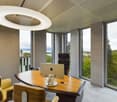Modern office space with large windows offering a scenic view, a curved wooden desk with an Apple laptop, and a circular lighting fixture above. The room has a minimalist and contemporary design with gray walls and a neutral color scheme.