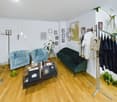 Living room with wooden floor, teal velvet chairs, green sofa, white walls, art on the wall, table with books and objects, clothes on a stand