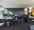 Modern office space with black and white furniture, multiple computer monitors, shelves with binders and folders, and a printer. The room has large windows providing natural light and a dark blue accent wall.