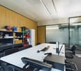 The image shows an office space with a large white desk, a computer, and various office supplies and equipment. The walls are lined with wooden panels, and there is a storage unit with shelves containing various items. The room also has large glass window
