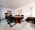The image shows a modern and minimalist office space. The room has white walls and a carpeted floor. There is a large oval-shaped wooden conference table in the center, surrounded by black leather chairs. On the walls, there is a framed abstract artwork a