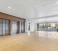 The image shows a large, open commercial space with a high ceiling, wooden wall panels, and glass doors leading to an outdoor area. The space has a modern, minimalist design with a polished tile floor and recessed lighting. There are two elevators visible