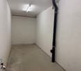 The image shows a small, empty storage room with concrete floors and white walls. There is a large pipe or conduit running along one of the walls.