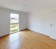 Spacious bedroom with hardwood flooring, white walls, and a large window providing natural light. The room appears to be empty, ready for furnishing and personalization.