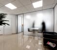 The image shows a modern office space with a tiled floor, white walls, and a suspended ceiling with recessed lighting. There is a potted plant in the corner, and a black office chair in the foreground. The room appears to be well-lit and has a clean, prof