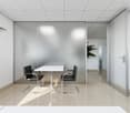 The image shows a modern and minimalist office space with a white table and two black chairs in the center. The walls are made of glass panels, providing a sense of openness and transparency. The ceiling is a grid-like pattern of perforated panels, creati
