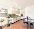 Modern, open-concept kitchen and dining area with light wood cabinetry, stainless steel appliances, and a dining table with black chairs. The space features large windows, potted plants, and a decorative stone wall accent.