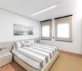 The image shows a modern and minimalist bedroom with a large bed, striped bedding, and a nightstand. The room has white walls, large windows, and a recessed lighting fixture on the ceiling. There is also a piece of abstract artwork hanging on the wall.