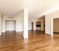 This image shows a spacious apartment interior with a large open floor plan. The room has hardwood floors, white walls, and a central column. There are multiple doorways leading to other rooms, including what appears to be a kitchen area. The overall desi