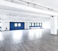 Large open commercial space with high ceilings, white walls, and gray hardwood flooring. There are blue garage doors at the far end of the room, indicating a garage or loading area.