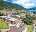 Large modern brick building with solar panels on the roof, surrounded by a lush green landscape and overlooking a lake and mountains in the background. The building appears to be situated in a picturesque town or village nestled in the mountains.