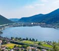 Aerial view of Riva San Vitale, a town located on the shores of a large lake surrounded by mountains. The town is situated along the lakefront with residential and commercial buildings, and there is a road running along the lake. The landscape is scenic w