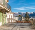 Facade of a building with balconies, green shutters, and tiled floor. A balcony with a metal railing. In the distance, mountains, trees, and buildings are visible.