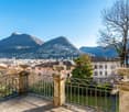balcony with metal railings, stone pedestals, large urns, metal fence, view of the city, mountains, and sea