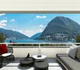 A covered balcony or terrace with modern furniture, offering a panoramic view of mountains and a city on a lake.