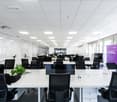 Large open office space with rows of desks and chairs, white walls, suspended ceiling with recessed lighting, and a purple banner on the wall that says 'Let there be change'.