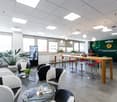 The image shows a modern and open office space with a mix of seating areas and workstations. The space features large windows, a high ceiling with recessed lighting, and a mix of furniture including glass tables, bar stools, and comfortable chairs. There
