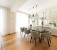 Dining room with wooden floor, large dining table, 6 chairs, pendant lights, white walls, white curtains, mirrors, plants
