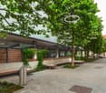 The image shows an outdoor seating area with benches and trees. The area appears to be part of a commercial or public space, with a paved walkway and buildings visible in the background. There is a parking area or space visible, indicating the presence of