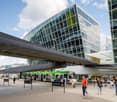 Large modern glass and steel commercial building with a covered bus station in the foreground. People are walking through the area, indicating it is a busy transportation hub.