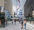 The image shows a modern commercial building with a glass facade and a covered walkway. Several people are walking through the walkway, which is lined with shops and businesses. The building has a sleek, contemporary design with a colorful, abstract sculp