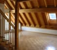 Spacious attic space with exposed wooden beams and rafters, hardwood flooring, and a window providing natural light.