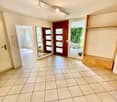 Empty room with beige tiles, white walls, wooden door, mirror, indoor plant, and window with balcony view.