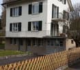 Two story house, brown roof, balcony, fenced garden, garages, white exterior, green windows, chimney, grass lawn, wooden fence