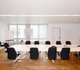 A modern, rectangular meeting room with white walls and wooden floors. The room features a large white table surrounded by black chairs. There is a whiteboard on an easel in the corner, and a whiteboard is mounted on the wall. There are windows with blind