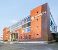 Exterior view of a modern office building with red brick walls and large glass windows. A clear blue sky is visible. The main feature is a large white and grey logo reading 'SPRICHS' on the front wall. The entrance has automatic glass doors and stairs. A
