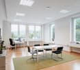 Empty office room with large windows, a desk, a meeting table with 4 chairs, a radiator, potted plants on the desk, and wooden floor.