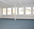 The image shows a large, open office space with white walls, a suspended ceiling, and large windows that provide natural light. The floor is covered in a gray, speckled carpet. The space appears to be empty and ready for use.