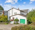 White house with brown roof, green garage door, brick driveway, garden, plants, shrubs, and trees