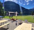 Outdoor area with tables and chairs, umbrellas, a tennis table, a wooden house in the background and a view of mountains.