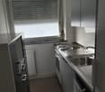 Stainless steel kitchen with white tiles, washing machine under counter, sink, and cooking stove, double radiator below window, drawers above washing machine, window blinds closed, metal drain above sink.