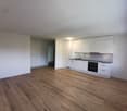 empty room with white walls, wooden floors, kitchen area with built-in appliances and white cabinetry