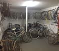 A bicycle storage area with bicycles hanging from hooks and placed on shelves, a white wall, and bicycles on the floor.
