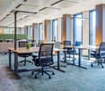 Spacious open-plan office with modern furniture and large windows providing natural light. The office features adjustable desks, ergonomic office chairs, and a green partition dividing the workspace. The flooring is a patterned carpet tile.