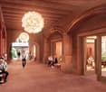 The image shows a grand, ornate interior space with high ceilings, arched doorways, and wooden beams. The space is illuminated by large, decorative chandeliers. Several people are visible, sitting and walking through the hallway. The overall atmosphere ap