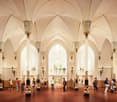 The image shows the interior of a large, ornate building with high ceilings and arched windows. The space is filled with people, and features decorative columns, chandeliers, and intricate architectural details. The floor is tiled in a checkered pattern.