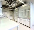 Modern laboratory with glass panels, white cabinets, white countertops, fluorescent lighting, glass shelves, and multiple control panels.
